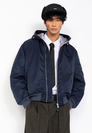 Navy bomber jacket features a zip front, ribbed cuffs and hem, and a hood. Details include a tag with branding and a smooth, shiny texture.
