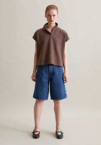 Braunes Polohemd mit kurzen Ärmeln und Kragen, kombiniert mit hellblauen knielangen Jeansshorts und schwarzen flachen Schuhen. Einfaches Design und klassischer Schnitt.