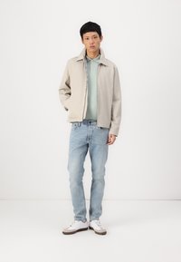 Veste bomber beige clair sur une chemise polo vert menthe, associée à un jean bleu clair et des baskets blanches avec des accents bruns.
