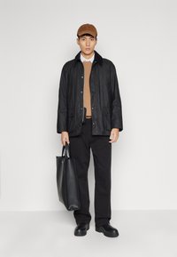 Zwarte waterdichte jas met een kraag, lichtbruine trui, zwarte broek, zwarte schoenen en een zwarte totebag. Een bruine pet maakt de look compleet.