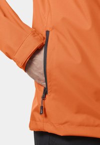 Oranžová bunda vyrobená z texturované tkaniny, s boční kapsou na zip a nastavitelnými manžetami. Zip má plastové táhlo.