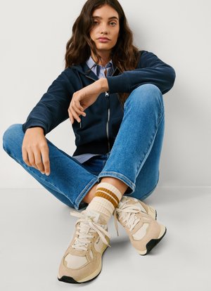 Marineblå jakke med glidelås, lyseblå skjorte, blå jeans, kremfargede joggesko med beige detaljer, og stripete kremfargede sokker med brune linjer.