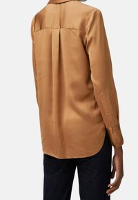 Camicia lunga in satin bronzo con colletto, dettagli con piega sul retro, polsini con bottoni e orlo curvo, abbinata a jeans scuri.
