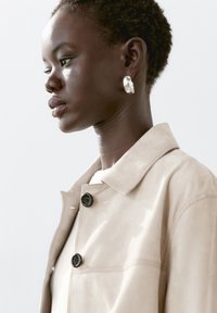 Beige mockasinjacka med krage, prydd med svarta knappar och en slät yta. Buren av en modell med kort hår och örhängen.