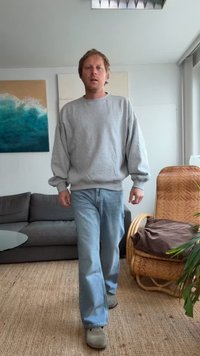 Sweatshirt cinza, jeans azul claro e sapatos cinza. Ambiente interno com um sofá cinza e uma cadeira de vime. Arte de parede com tons azul-esverdeados.
