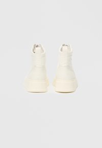 Par av høyhælte, off-white sneakers vist fra baksiden på en ensfarget lys grå bakgrunn.