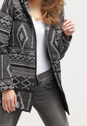 Vrouw die een zwart-grijze jacquet met patroon draagt over een wit top en grijze jeans, poseert met één hand omhoog bij de borst.