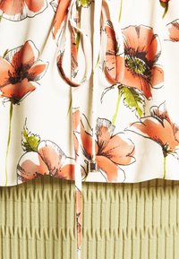 Kwiatowa bluzka z dużymi pomarańczowymi wzorami maków na białym tle, z wiązaniem z metalowym akcentem, nad żeberkowym zielonym materiałem.