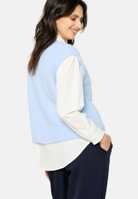 Gaiši zila flīsa veste ar mīkstu tekstūru, ar brīvu piegriezumu un bez piedurknēm, uzvilkta pār balto krekli.