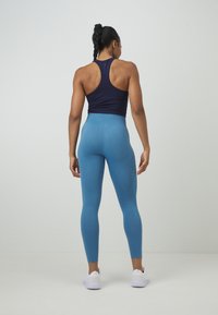Top in nylon blu navy con design a racerback, abbinato a leggings blu a vita alta con texture e scarpe da ginnastica bianche. Vista posteriore mostrata.