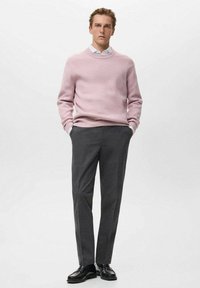 Jasnoróżowy sweter prążkowany noszony na białej koszuli, w zestawie z ciemnoszarymi spodniami w kant i czarnymi skórzanymi butami. Model stoi na jasnym tle.