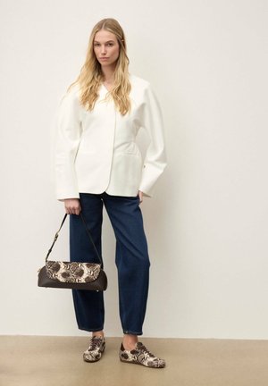 Femme portant un blazer blanc, un jean bleu et des baskets imprimé serpent, tenant un sac à main assorti à imprimé serpent sur un fond uni.