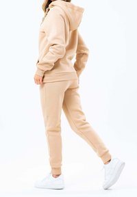 Béžová mikina s kapucí a odpovídající tepláková souprava z měkké látky. Má kapuci, elastické manžety a bílé tenisky. Hladká textura, jednoduchý design.