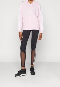 Svetle růžová mikina na zip s bílými pruhy, v kombinaci s černými legínami s síťovými akcenty a bočními pruhy. Černé sportovní boty dokončují outfit.