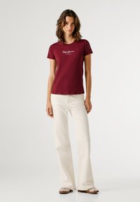 Mladá žena stojí v bordovej tričku s krátkym rukávom a bielym textom, krémových širokých nohaviciach a bielych vzorovaných topánkach.