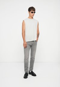 Top grigio senza maniche, jeans slim-fit grigio chiaro e mocassini neri. Gli accessori includono occhiali da sole scuri. Texture dei tessuti morbide.