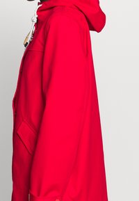 Chaqueta roja con capucha de textura suave, que cuenta con un bolsillo lateral y un cierre con cordón. El diseño es sencillo y funcional.