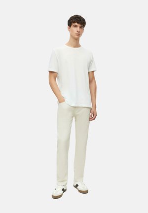 Junger Mann steht und trägt ein einfaches weißes Kurzarm-T-Shirt, hellbeige Hosen und weiße Sneaker mit schwarzen Akzenten.
