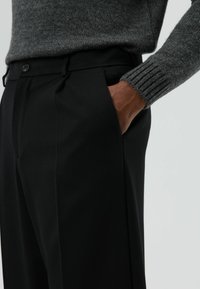 Zwarte broek met een getailleerde pasvorm, voorzien van voorplooien, een knoopsluiting en zijzakken. Gecombineerd met een donkergrijze textuurtrui.