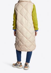 Person, der eine lange beige gesteppte Weste über einem grünen Pullover, blauen Jeans und mehrfarbigen klobigen Turnschuhen trägt, mit dem Rücken zur Kamera auf weißem Hintergrund.