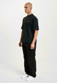 Black t-shirt with short sleeves and a crew neck, paired with black cargo pants and white sneakers, worn by a person standing sideways.