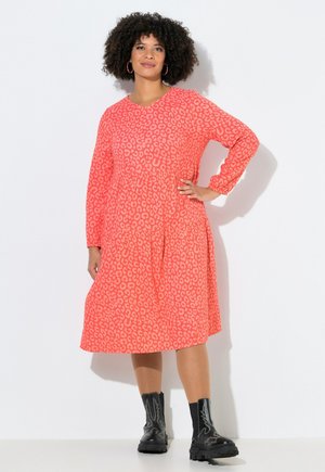 Kvinde med krøllet hår iført en koral pink leopard print langærmet kjole og sorte støvler til midt på læggen, står med hånden på hoften mod en hvid baggrund.