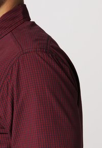Gros plan d’une personne portant une chemise rouge et noire à carreaux à manches longues sur un fond clair uni.