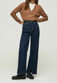 Brązowy sweter z dekoltem w serek, biały podkoszulek, ciemnoniebieskie szerokie dżinsy z wysokim stanem i rozcięciami z przodu, czarne masywne botki na obcasie.