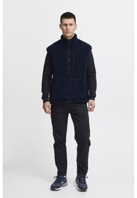 Mornarska flis prsluk s visokim ovratnikom, zatvaračem sprijeda i dvjema džepovima. Nosite ga preko crne košulje dugih rukava, u kombinaciji s crnim hlačama i tenisicama.