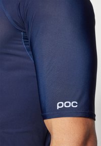 Mornarice plava sportska majica uskog kroja, s kratkim rukavima i malim bijelim "poc" logotipom blizu manžeta, izrađena od glatkog, rastezljivog materijala.