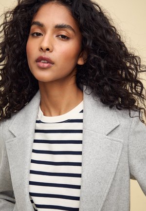 Blazer en laine gris avec des revers en pointe sur une chemise rayée crème et marine. Le tissu semble doux. Les cheveux sont bouclés et de longueur épaule.