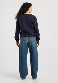 Mujer con cabello rizado que lleva un sudadera azul marino y jeans azules holgados, de pie de espaldas contra un fondo claro y liso.