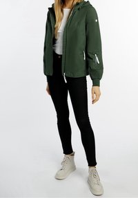 Veste à capuche vert foncé avec poches zippées, poignets élastiques et accessoires blancs, associée à un jean skinny noir et des baskets blanches.