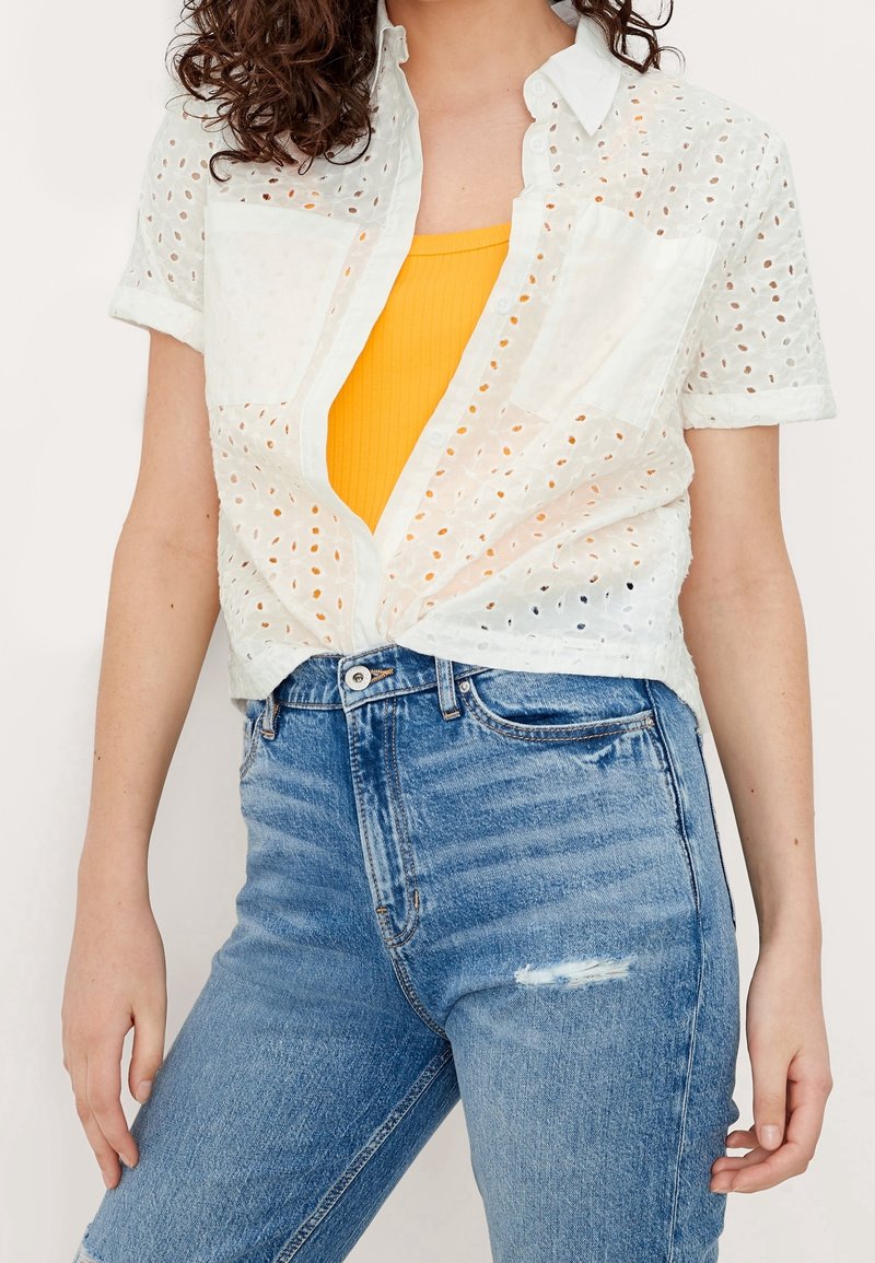 Femme portant une chemise blanche à manches courtes en œillet transparente, boutonnée, sur un débardeur jaune et un jean en denim bleu déchiré.