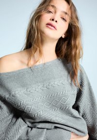 Pulover gri tricotat, cu textură canelată, design off-shoulder și modele complicate de cablu pe întreaga suprafață. Material moale, casual.