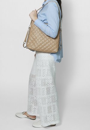 Frau trägt ein blaues Hemd und weiße, weit geschnittene Spitzenhosen, dazu eine große, gesteppt beige Schultertasche mit Kettendetails.