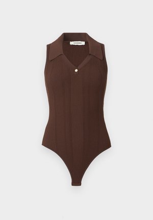 Γυναίκα στέκεται φορώντας αμάνικο καφέ ριμπ πουκάμισο με γιακά, τζιν παντελόνι με ψηλή μέση και μεγάλες εμπρόσθιες τσέπες και μαύρα μοκασίνια.