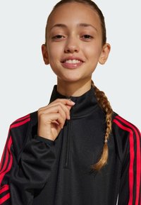 Schwarze Sportjacke mit hohem Kragen, ausgestattet mit roten Streifen auf den Schultern und einem Halben Reißverschluss. Glatte Textur, taillierte Passform.