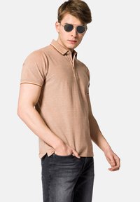 Beżowa koszulka polo z klasycznym kołnierzykiem, z kontrastowymi prążkowanymi brzegami. Noszona z ciemnymi dżinsami, uzupełniona okrągłymi okularami przeciwsłonecznymi.