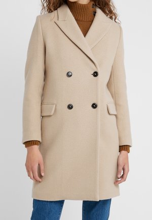 Cappotto in lana doppiopetto beige con bottoni neri, rever a taglio, tasche laterali e maglione marrone a coste visibile sotto.