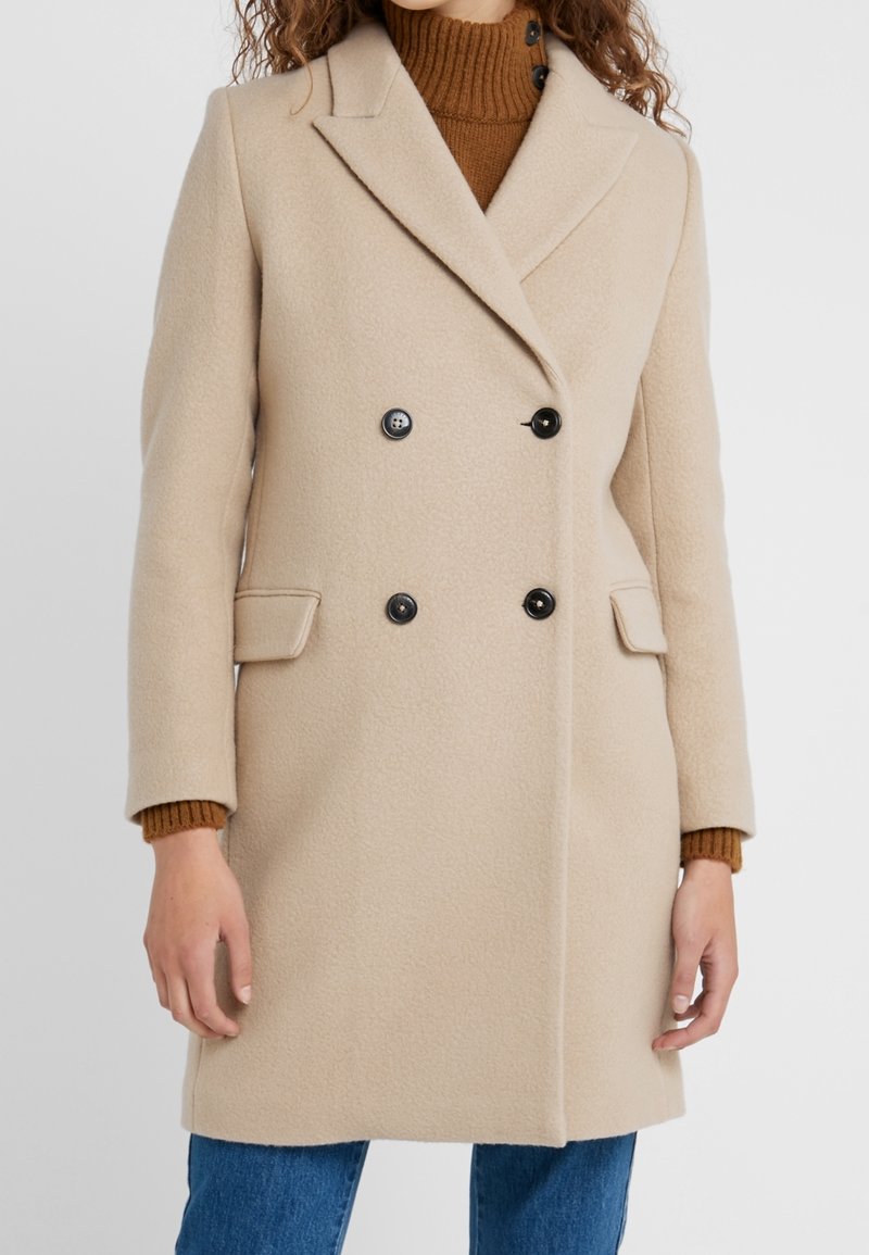 Beige Doppelreiher-Wollmantel mit schwarzen Knöpfen, Reverskragen, seitlichen Taschen und unter dem Mantel sichtbarem braunem Rippenpullover.