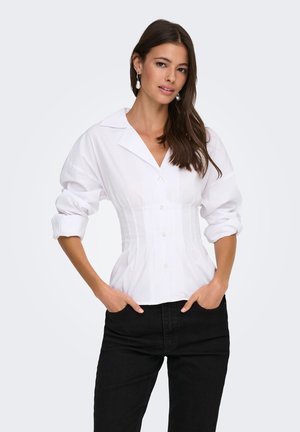 Femme aux longs cheveux foncés portant un chemisier blanc ajusté boutonné avec manches retroussées et un pantalon noir, debout les mains dans les poches.
