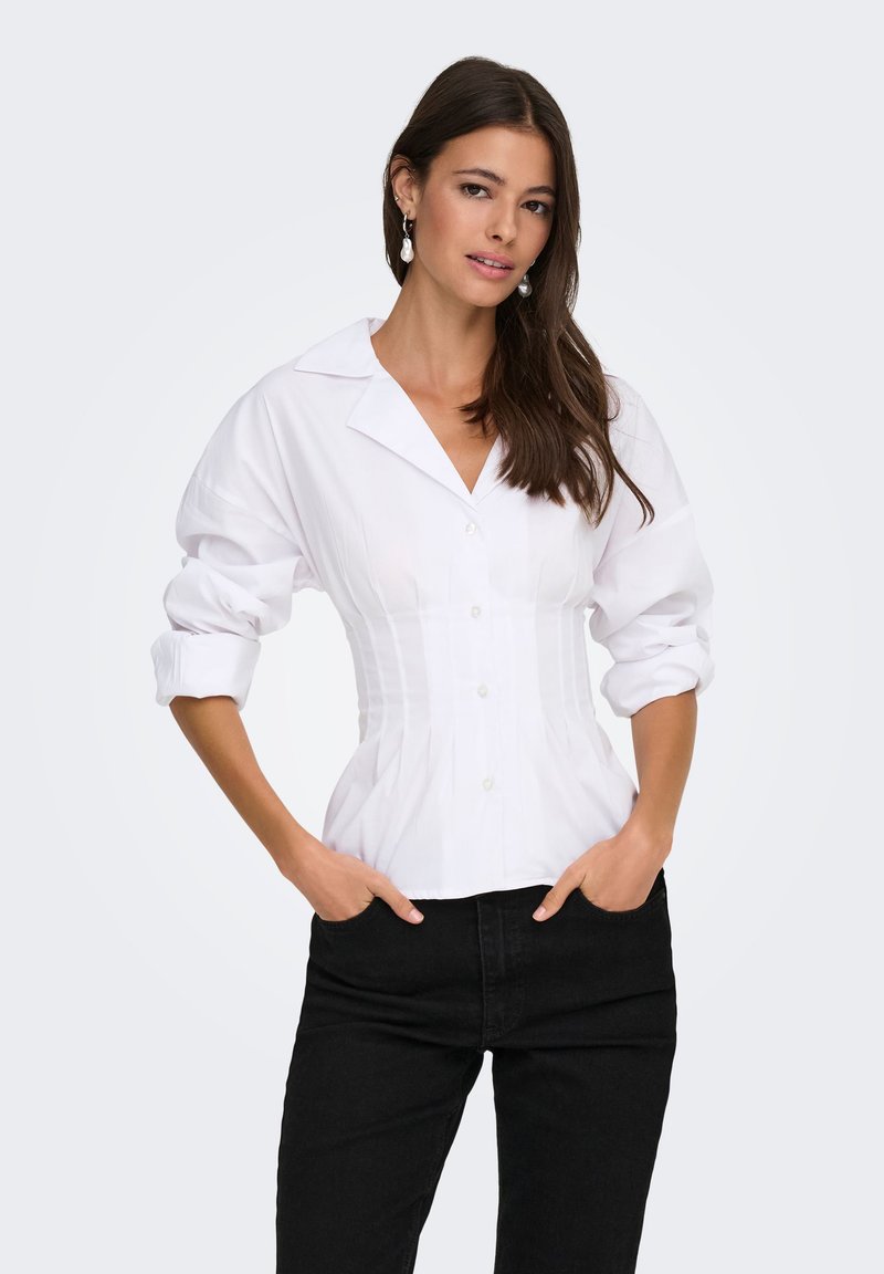 Femme aux longs cheveux foncés portant un chemisier blanc ajusté boutonné avec manches retroussées et un pantalon noir, debout les mains dans les poches.