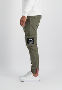 Olivenfarbene Cargo-Hosen mit elastischen Bündchen, mehreren Taschen und einem schwarzen Etikett mit weißem Text. Das Material scheint leicht und langlebig zu sein.