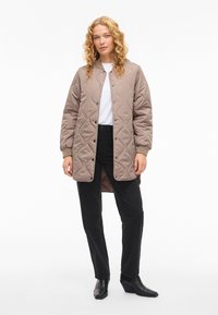 Beżowa pikowana kurtka z zapięciem na zatrzaski, ściągaczami przy rękawach oraz lekko wydłużonym tyłem, w zestawieniu z czarnymi spodniami i botkami za kostkę.