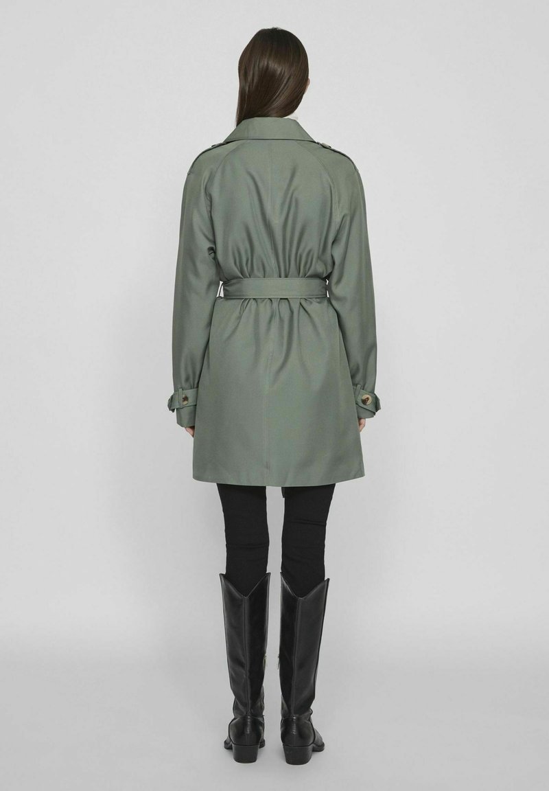 Olijfgroen trenchcoat met een ceintuur in de taille, lange mouwen en knopenmanchetten. Gestyled met zwarte strakke broeken en zwarte laarzen tot aan de knie.