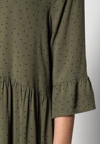 Olivově zelené šaty s rukávem, pokryté černými puntíky, s detailem nařaseného švu a částečně viditelnou odhalenou paží.