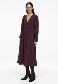 Langärmliges Kleid mit tiefem V-Ausschnitt aus dunkelviolettem Stoff, vertikalen Falten und Seitentaschen, kombiniert mit kniehohen schwarzen High Heels Stiefeln.