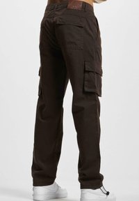 Pantalons cargo marron en coton, dotés de plusieurs poches latérales, d'une patch en cuir avec "DEF" et d'une coupe décontractée. Des chaussures blanches visibles en bas.