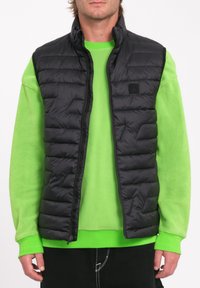 Il gilet a puffo nero presenta una chiusura con zip, cuciture orizzontali e tasche laterali. Indossato sopra una felpa in pile verde brillante con polsini a coste.